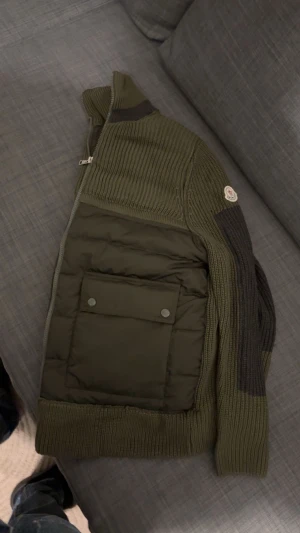 Unik olivgrön Moncler cardigan jacka - Olivgrön jacka från Moncler med stickade ärmar och pufferfront. Jackan har två stora fickor. Perfekt för dig som vill ha en stil med premiumkänsla. Har även NFC scan.