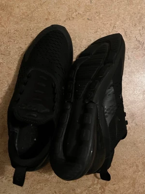 Svarta Nike Air Max 270 sneakers - Säljer ett par helsvarta Nike Air Max 270 sneakers med mesh-ovandel och synlig Air-enhet i hälen. Skorna har en sportig look med diskret Swoosh-logga på sidan och dragflik bak. Perfekta för dig som gillar stilrena och bekväma sneakers.
