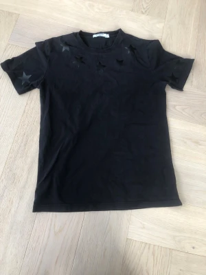 Svart t-shirt med stjärnor från Givenchy - Snygg svart t-shirt från Givenchy med mattsvarta stjärnor längs axlarna och på ärmarna. Klassisk rund halsringning och korta ärmar. Tillverkad i mjuk bomull för en skön känsla och stilren look.