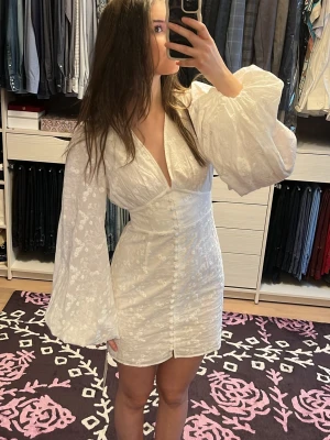 Vit blommig klänning från Adoore - Supersöt vit klänning från Adoore med blommigt broderat mönster, v-ringning och dekorativa knappar framtill. Klänningen har långa puffärmar och är tillverkad i ett lätt tyg som känns lyxigt. Perfekt för sommarens alla tillfällen SÄRSKILLT STUDENTEN🥳🎓!  Aldrig använd, helt ny med ettiketter 