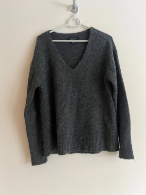 Mörkgrå stickad tröja med V-ringning - Mysig mörkgrå stickad tröja från Aylin med djup V-ringning och långa ärmar. Tröjan har ribbstickade muddar vid ärmslut och nederkant. Perfekt för lager på lager och enkel att matcha till olika outfits.