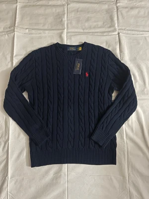 Mörkblå kabelstickad tröja Polo Ralph Lauren - Snygg mörkblå kabelstickad tröja från Polo Ralph Lauren med rund halsringning och den klassiska röda logotypen broderad på bröstet. Tröjan har långa ärmar och ribbade muddar. Perfekt för dig som vill ha en tidlös och stilren look.