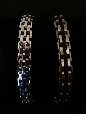 Två kedjearmband i metall - Säljer två stilrena armband i silverfärgad metall med bred kedjedesign. Ena armbandet har svarta detaljer mellan länkarna, medan det andra är helt i silver. Perfekta för dig som gillar en modern och edgy look.