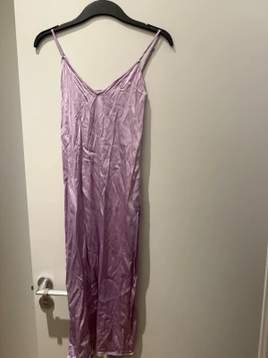Ljuslila satin klänning med smala band - Säljer en lång klänning i ljuslila satin med smala axelband och v-ringning fram. Materialet är glansigt och mjukt, vilket ger en lyxig känsla. Perfekt för mysiga kvällar hemma. Enkel och stilren design utan mönster.