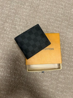 Louis Vuitton svart rutmönstrad plånbok - Snygg svart plånbok från Louis Vuitton med klassiskt rutmönster i canvas. Plånboken har en stilren och kompakt design, perfekt för kort och sedlar. Levereras med original ask och dustbag.