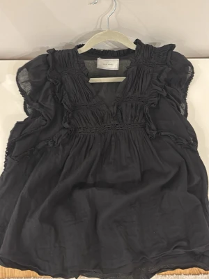 Svart volangblus Neo Noir - Snygg svart blus från Neo Noir med volangdetaljer och v-ringning. Blusen har korta ärmar och är tillverkad i ett tunt, luftigt tyg som ger en avslappnad men trendig look. Perfekt att styla med jeans eller kjol för en cool outfit.