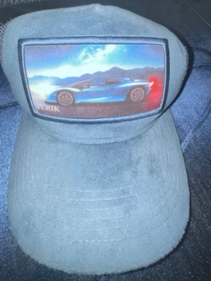 Blå truckerkeps med bilmotiv - Säljer en blå truckerkeps från PERIK med mesh på sidorna och ett stort tygmärke framtill med motiv av en blå sportbil mot berg och himmel. Kepsen har böjd skärm och justerbar baksida. Perfekt för dig som gillar bilar och streetwear.