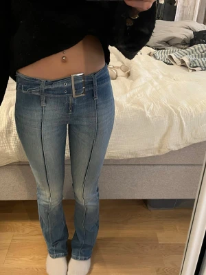 Blå jeans med markerad söm och bälte - Säljer ett par ljusblå jeans med raka ben och markerad pressveckssöm framtill. Jeansen har ett brett midjebälte med stor silverfärgad spänne som detalj. Perfekta för en trendig och avslappnad look.
