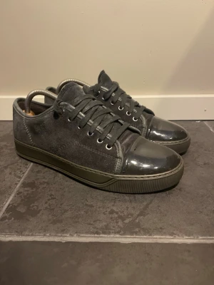 Mörkgrå sneakers med lackdetaljer - Snygga mörkgrå sneakers med ovandel i mocka och glansiga lackdetaljer på tån och hälen. Skorna har matchande mörkgrå snören och en låg silhuett som ger en clean look. Perfekta för dig som vill ha något stilrent men ändå unikt.