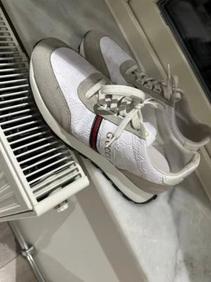 Vita sneakers med beige detaljer - Snygga vita sneakers med beige mockadetaljer och klassiska gröna och röda ränder på sidan. Skorna har präglat GG-mönster i tyget och guldfärgad logga på hälen. Låga i modellen och med vita snören. Storlek 41