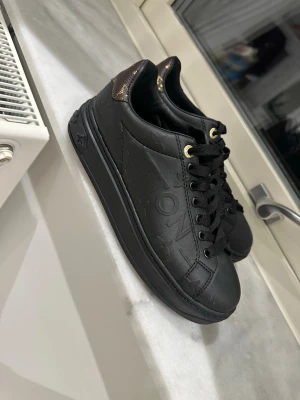 Svarta  sneakers - Snygga svarta sneakers Louis Vuitton i läder med präglad logga på sidan och guldfärgade detaljer vid snörningen. Skorna har klassisk låg profil och svart sula. Perfekta för dig som gillar exklusiv streetstyle.