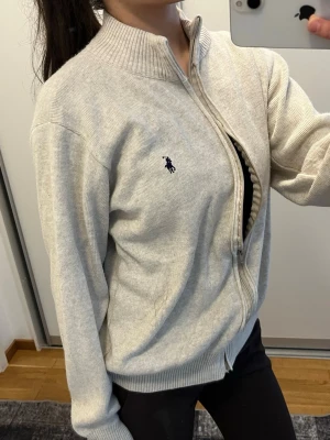 Ljusgrå kofta från Ralph Lauren - Ljusgrå ziptröja från Ralph Lauren med hög krage och ribbade muddar. Tröjan har det klassiska broderade logotypen på bröstet och är tillverkad i mjukt stickat material. Perfekt för lager på lager och enkel att matcha med olika outfits.