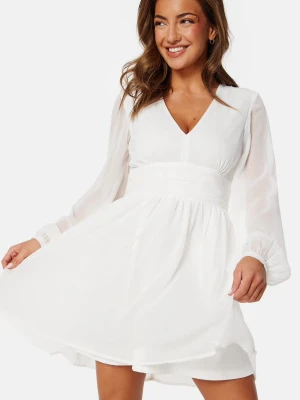 Vit kortklänning med v-ringning - Vit kortklänning med v-ringning och långa, transparenta ärmar. Klänningen har markerad midja. Perfekt för sommarens alla tillfällen och studenten.