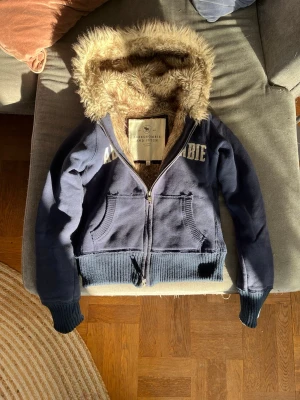 Blå hoodie från Abercrombie & Fitch - Mysig blå hoodie med dragkedja från Abercrombie & Fitch. Stor huva fodrad med mjuk, beige fuskpäls och ribbade muddar vid ärmar och midja. Stora fickor framtill och tryckt logga på bröstet. Perfekt för dig som gillar en avslappnad och trendig stil.