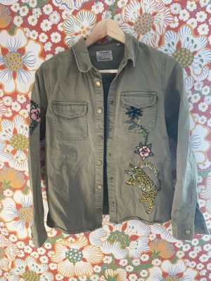 Grön Zadig skjorta med broderier och leopard - Stilren grön skjorta med broderade blommor och en leoparddetalj på framsidan. Två bröstfickor med lock och knappar framtill. Skjortan har lång ärm och en avslappnad passform. Perfekt för dig som vill sticka ut lite extra. Liten fläck vid knapparna.