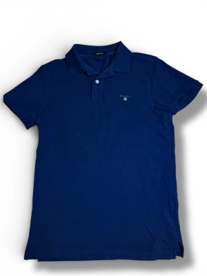 Marinblå pikétröja från Gant - Snygg marinblå pikétröja från Gant med klassisk krage och korta ärmar. Tröjan har två knappar vid halsen och en liten broderad logotyp på bröstet. Perfekt för en stilren och avslappnad look.