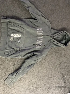 Grå hoodie från Hoodrich med tryck - Snygg grå hoodie från Hoodrich med stor logga på bröstet och tryck på magfickan. Klassisk känguruficka och justerbar huva med snören. Tillverkad i mjukt bomullsmaterial som är skönt mot huden. Perfekt för en avslappnad streetwear-look.