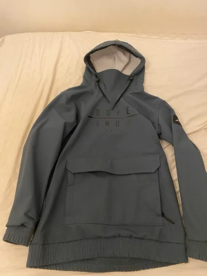 Blå anorak från Dope Snow - Säljer en stilren blå anorak från Dope Snow med stor magficka, huva med dragsko och diskret logga på bröstet. Jackan har elastiska muddar och är tillverkad i ett slitstarkt material som passar perfekt för vintersport och utomhusaktiviteter.