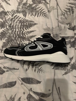 Dior b30 - Säljer ett par svarta Dior sneakers med vita och grå detaljer samt vita skosnören. Skorna har en chunky vit sula och tydliga Dior-loggor på sidan och plösen. Materialet är en mix av mocka och mesh för en sportig och exklusiv look. Perfekta för dig som vill sticka ut.