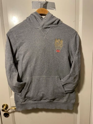 Grå hoodie från morris - Grå hoodie från Morris med broderad guldfärgad logga och liten brittisk flagga på bröstet. Klassisk känguruficka framtill och ribbade muddar. Perfekt för en avslappnad stil.