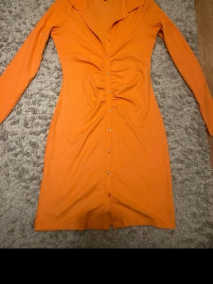 Orange ribbad kortklänning - Snygg orange kortklänning med ribbad struktur och långa ärmar. Klänningen har knappar framtill, v-ringning och en smickrande rynkning längs midjan. Perfekt för dig som vill sticka ut med färg och stil.