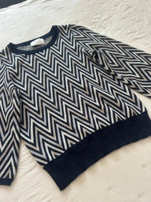 Svartvit zigzag-stickad tröja från Ahlvar - Snygg stickad tröja från neo noir i ett marinblått, beiget och vitt mönster, tröjan har trekvartsärmar och en mudd nertill som  ger en snygg passform. 
