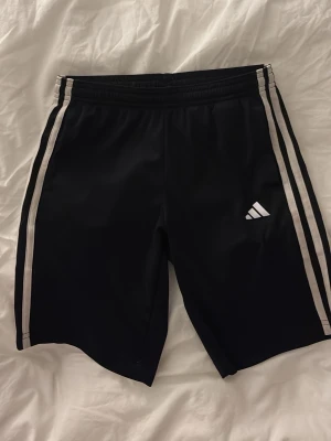 Svarta Adidas shorts med vita ränder - Säljer ett par svarta Adidas shorts med klassiska vita ränder längs sidorna och Adidas-logga på ena benet. Shortsen har elastisk midja och är tillverkade i ett lätt och bekvämt material som passar perfekt till sport och träning. storlek 9-10 år men passar xs-s!!