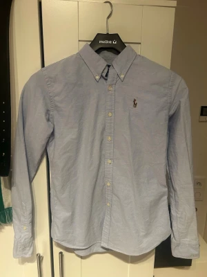 Ljusblå skjorta från Ralph Lauren - Klassisk ljusblå skjorta från Ralph Lauren med button-down krage och broderad logga på bröstet. Skjortan har långa ärmar, knappar framtill och är tillverkad i bomull. Perfekt för en clean och stilren look. 