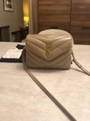 Beige YSL axelväska i läder - Säljer min Saint Laurent Loulou Toy i färgen dusty beige med guld detaljer 🤍  Väskan är i superfint skick utvändigt. Insidan är i svart tyg och det kan finnas några små sminkmärken.  Den är smidig i storleken och kan bäras både på axeln och crossbody   Originalkvitto från Saint Laurent-butiken på NK i Göteborg samt dustbag medföljer 🤍