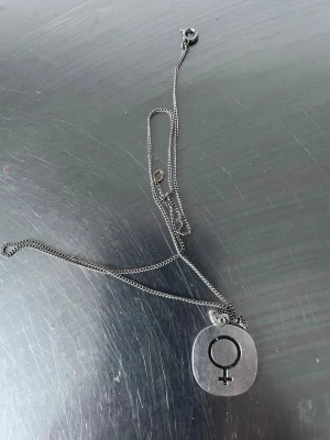 Silverfärgat halsband med kvinnosymbol - Snyggt halsband i silverfärgad metall med en rundad, platt berlock som har en ingraverad kvinnosymbol. Kedjan är tunn och har ett klassiskt lås. Perfekt accessoar för att uttrycka din stil och personlighet.