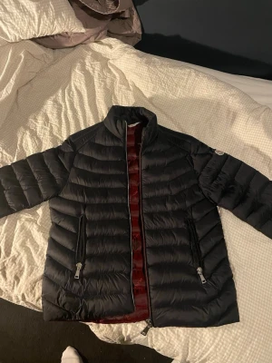 Svart dunjacka med röd insida - Snygg svart dunjacka med quiltad design och hög krage. Jackan har två dragkedjeförsedda fickor framtill och en cool röd insida. Perfekt för kalla vinterdagar och riktigt lätt att bära. Passar dig som vill ha både stil och värme.