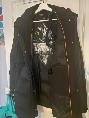 Svart lång dunjacka från Hollies - Snygg svart lång dunjacka från Hollies med huva och dragkedja framtill. Jackan har orange detaljer på insidan och trycket 'Arctic Expedition' på fodret. Två framfickor med dragkedja och quiltad design för extra värme. Perfekt för kalla vinterdagar. En knapp saknas bak 