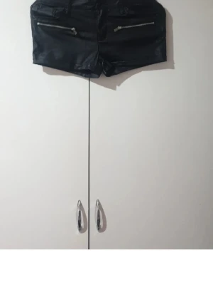 Svarta shorts i skinnimitation - Snygga svarta shorts i skinnimitation med två dragkedjor framtill och fickor både fram och bak. De har en tight passform och korta ben, perfekta för en edgy stil. Passar till många olika looks och är lätta att matcha.