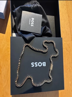 Kedja i stål från BOSS - Stilren kedja från BOSS i metallisk, silverfärgad finish. Kedjan har en fyrkantig, modern design och ett diskret BOSS-märke på låset. Kommer i originalförpackning med svart tygpåse och etikett. Perfekt accessoar för dig som gillar minimalistiska och trendiga detaljer.