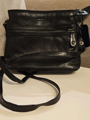 Svart axelväska i skinn från Coach - Snygg svart axelväska från Coach i äkta skinn med justerbar rem. Väskan har flera fack med dragkedja och en stilren, klassisk design. Perfekt för dig som vill ha en praktisk men ändå trendig väska med plats för det viktigaste.