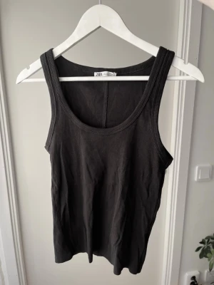 Svart ribbad linne topp från Zara - Säljer en svart ribbad linnetopp från Zara. Toppen har breda axelband och rundad halsringning. Perfekt att styla lager på lager eller bära som den är. Materialet är mjukt och stretchigt för en bekväm passform.