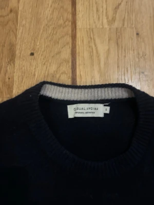 Mörkblå stickad tröja Casual Aesthetics - Mörkblå stickad tröja från Casual Aesthetics med rund halsringning och diskret ljusgrå kant på insidan av kragen. Klassisk modell med långa ärmar och ribbstickade muddar. Perfekt för en stilren och enkel look. Nypris 799