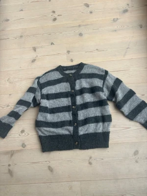 Stickad randig kofta  - Mysig stickad kofta med breda horisontella ränder i mörkgrått och ljusgrått. Koftan har rund halsringning, knappar framtill och ribbstickade muddar vid ärmslut och nederkant. Så fin och mjuk!