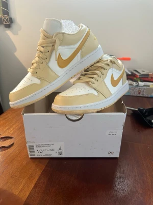 Nike Air Jordan 1 Low beige/vit - Snygga Nike Air Jordan 1 Low sneakers i beige och vitt med klassisk guldfärgad swoosh. Skorna har en låg silhuett, perforerad tåbox och ovandel i läder. Perfekta för dig som gillar streetwear och vill sticka ut med stilrena detaljer.