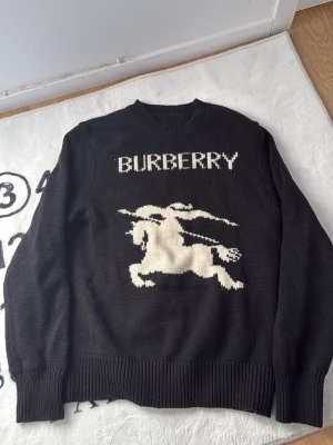 Svart stickad Burberry-tröja - Stickad svart tröja från Burberry med vit logotyp och en stor riddare på häst framtill. Tröjan har rund halsringning, ribbade muddar och är gjord i ett mjukt material som känns lyxigt. Perfekt för dig som vill ha något stilrent men ändå med statement.