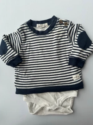 Randig body med lång ärm från Newbie 62 - Söt långärmad body från Newbie i marinblått och vitt med smala ränder. Bodyn har mörkblå muddar, knappar på axeln och förstärkta armbågar. Tillverkad i mjuk bomull, perfekt för en bekväm och stilren look.