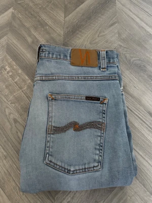 Ljusblå lean dean från Nudie Jeans - Säljer ett par ljusblå lean dean jeans från Nudie Jeans med klassisk femficksmodell och snygga slitningar. Jeansen har en broderad bakficka och patch i läder bak i midjan. Tillverkade i mjuk denim, perfekta för en avslappnad stil. Priset kan diskuteras 