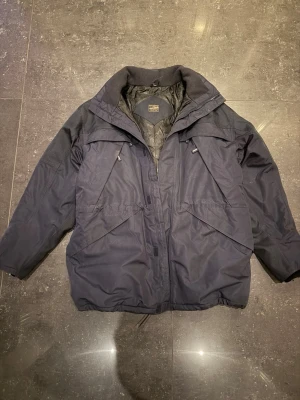 Mörkblå vindjacka från Fristads  - Snygg mörkblå vindjacka från Fristads med quiltat foder och hög krage och 11 fickor. Jackan har dragkedja framtill, flera praktiska fickor och justerbar midja. Materialet är slitstarkt och vattenavvisande, perfekt för blåsiga dagar. Stilsäker och funktionell design. 