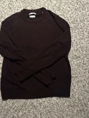 Mörkbrun stickad tröja These Glory Days - Mörkbrun stickad tröja från These Glory Days med rund halsringning och ribbade muddar. Enkel och stilren design som passar perfekt till jeans eller chinos. Tillverkad i mjukt material för en bekväm känsla.