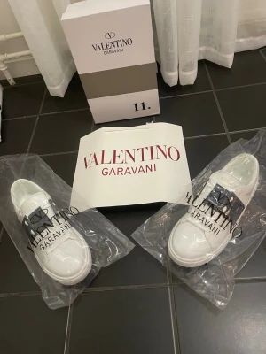 Vita Valentino Garavani sneakers - Snygga vita sneakers från Valentino Garavani med svart detalj över snörningen. Klassisk låg modell med rund tå och tjock sula. Tillverkade i läder för en lyxig känsla och stilren look. Perfekta för dig som vill ha något exklusivt och trendigt.