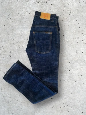Mörkblå jeans från Nudie Jeans - Säljer ett par mörkblå jeans från Nudie Jeans med klassisk femficksdesign och kontrastsömmar. Modellen är Thin Finn. Jeansen har rak passform och är tillverkade i kraftig denim. Perfekta för dig som gillar stilrena och tidlösa jeans.
