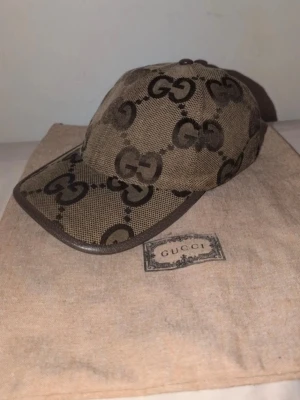 Beige Gucci jumbo keps  - Snygg beige keps från Gucci med det klassiska GG-monogrammet i brunt över hela kepsen. Kepsen har böjd skärm, justerbar rem bak och är tillverkad i canvasmaterial med läderdetaljer. Perfekt för dig som gillar exklusiva accessoarer och streetstyle.