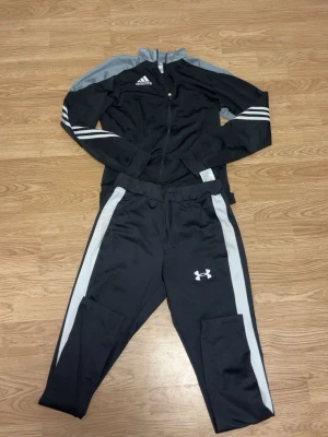 Svart träningsset Adidas & Under Armour - Snyggt svart träningsset med Adidas ziptröja och Under Armour byxor. Tröjan har vita ränder på ärmarna och grå detaljer på axlarna. Byxorna har vita sidopaneler och Under Armour-logga på benet. Perfekt för gymmet eller löprundan.