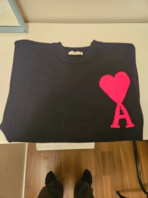  Ami Paris stickad tröja - Säljer en svart stickad tröja från Ami Paris med deras ikoniska röda hjärta och bokstaven 'A' broderat på bröstet. Tröjan har rund hals, långa ärmar och ribbade muddar vid ärmslut och nederkant. Perfekt för dig som gillar stilrena detaljer.