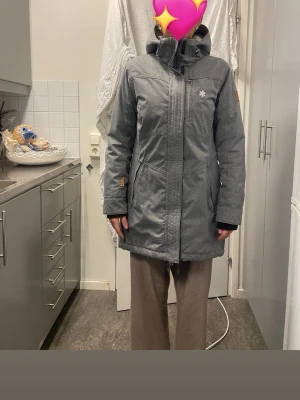 Grå parkasjacka från Ahkka - Snygg grå parkasjacka från Ahkkamed hög krage och stor huva. Jackan har dragkedja och vindslå framtill, samt diskreta loggor på bröstet och ryggen. Praktiska fickor med dragkedja och justerbara ärmslut. Perfekt för kalla dagar.